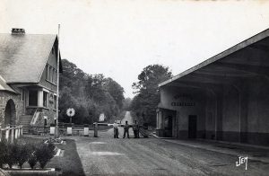 Poste-frontière ancien entre la France et la Belgique en Thiérache. Au centre de la scène, trois douaniers se tiennent devant une barrière en bois qui coupe la route. À droite, un grand bâtiment surélevé porte l’inscription « Douanes Françaises ». À gauche, une maison en pierre au style régional domine légèrement la chaussée. La route rectiligne file vers une forêt dense, plongeant dans un paysage rural et boisé. L’ensemble évoque une atmosphère calme, typique des postes douaniers d’autrefois.
