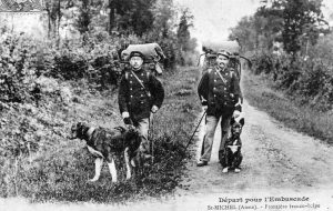 Deux douaniers français en uniforme, sacs à dos chargés, marchent avec leurs chiens le long d’un chemin bordé de végétation. La scène, en noir et blanc, date du début du XXᵉ siècle. L’inscription en bas de l’image indique : “Départ pour l’embuscade – St-Michel (Aisne), frontière franco-belge