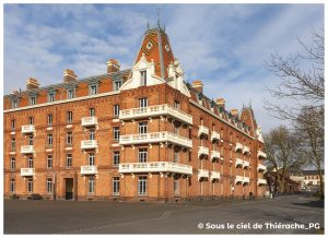 Aile du Palais Social du Familistère de Guise vue depuis la place, grand bâtiment en briques rouges avec balcons blancs, toitures ornées et lucarnes dans un ciel clair d’hiver