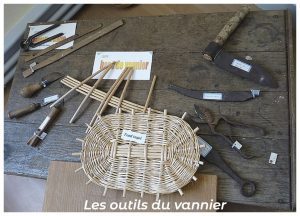 able présentant différents outils traditionnels de vannier — serpettes, couteaux, poinçons, ciseaux, gabarits en bois — accompagnés d’un fond de panier en osier en cours de fabrication, illustrant les instruments essentiels du tressage artisanal.