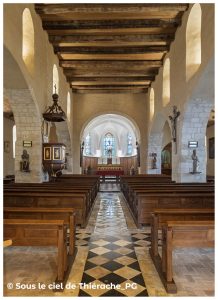Intérieur de l’église Notre-Dame de Plomion : vue centrale vers le chœur, avec nef aux murs en pierre claire, plafond en poutres de bois massif, rangées de bancs en chêne et dallage géométrique en pierre. Au fond, autel éclairé par trois vitraux colorés. Ambiance sobre et lumineuse du patrimoine thiérachien.