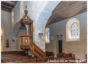 Chaire en bois sculpté et doré de l’église Notre-Dame de Plomion, posée contre un pilier en pierre claire. L’escalier et la cuve présentent des ornements dorés. À droite, deux vitraux colorés éclairent la nef. Bancs en bois et plafond lambrissé complètent l’ambiance chaleureuse de l’intérieur thiérachien.