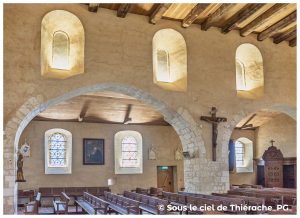 ntérieur de l’église Notre-Dame de Plomion, montrant les arcades en pierre, les murs enduits couleur sable et les petites fenêtres romanes laissant entrer une lumière douce. Des bancs en bois occupent la nef, tandis que des vitraux colorés illuminent l’espace. Un grand crucifix en bois est fixé contre un pilier, et le plafond en bois massif renforce l’atmosphère chaleureuse de cet intérieur thiérachien.