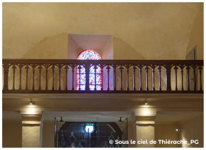 Tribune en bois sculpté de l’église Notre-Dame de Plomion, éclairée par un vitrail aux couleurs vives en arrière-plan. La balustrade gothique se détache sur les murs enduits aux tons chauds. En dessous, l’espace d’entrée avec éclairage doux et porte vitrée moderne s’intègre au patrimoine thiérachien.