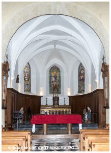 Chœur de l’église Notre-Dame de Plomion : voûtes gothiques blanches, vitraux colorés diffusant une lumière douce, autel orné de boiseries sculptées et d’un retable décoré. À l’avant, une table couverte d’un drap rouge contraste avec les bancs en bois clair et le dallage géométrique typique du patrimoine thiérachien.