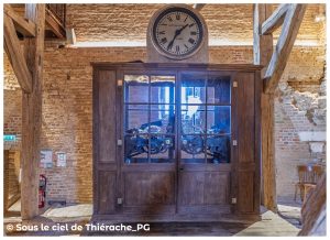 Ancien mécanisme d’horloge de l’église Notre-Dame de Plomion, présenté dans une armoire vitrée en bois. À son sommet, un cadran rond marqué « Leroy & Fils, Paris ». À l’intérieur, engrenages et rouages métalliques mis en valeur par un éclairage bleu. L’ensemble est installé dans les combles en briques et poutres anciennes du bâtiment.