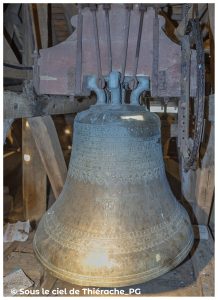 Cloche ancienne du clocher de l’église Notre-Dame de Plomion : grande cloche en bronze ornée de frises et d’inscriptions, suspendue à une structure en bois et métal patiné. On distingue un Christ crucifié gravé sur la panse. L’ensemble est installé dans la charpente sombre du clocher, éclairé par une lumière rasante révélant la texture du métal.