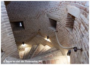 Escalier en vis de la tour gauche de l’église Notre-Dame de Plomion : marches en pierre usées, murs circulaires en briques appareillées et petites meurtrières laissant filtrer la lumière. Une corde fixée au mur sert de main courante, éclairée par des appliques discrètes qui révèlent la structure défensive du bâtiment.