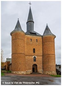 Façade monumentale en briques de l’église fortifiée Notre-Dame de Plomion, avec son donjon central carré et ses deux puissantes tours rondes coiffées de toits en poivrière. Le clocher élancé domine l’ensemble, tandis que l’entrée s’ouvre au pied du massif défensif. Sol humide et ciel gris typiques d’un paysage thiérachien.