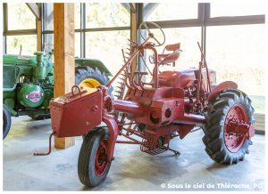 Engin agricole ancien de couleur rouge exposé dans un musée. L’engin, au design atypique avec un siège en hauteur, un grand volant et des éléments mécaniques apparents, est présenté sur un sol en béton près de grandes baies vitrées. À l’arrière-plan, on aperçoit un autre tracteur vert portant l’inscription « Société Française Vierzon ». L’ensemble évoque une collection de machines agricoles historiques.