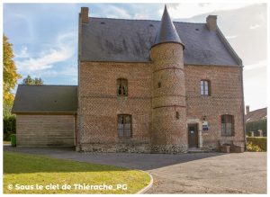 Façade en briques rouges de la Maison du Prévôt à Saint-Michel, surmontée d’un haut toit d’ardoises. Une tourelle cylindrique avec toiture conique s’élève au centre de la façade, encadrée par des fenêtres aux arcs de briques. À droite, une porte ancienne porte l’inscription « Maison du Prévôt ». À gauche, une extension en bois complète le bâtiment. L’ensemble est éclairé par un soleil doux, avec un ciel bleu légèrement nuageux et un gazon au premier plan.