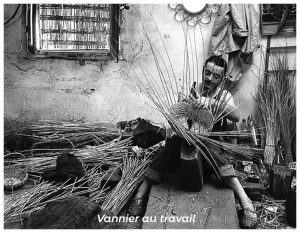 Artisan vannier assis dans son atelier, en train de tresser un panier en osier, entouré de fagots de brins végétaux et d’outils, scène capturant la concentration et le geste traditionnel du métier.