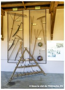 Panneau d’exposition consacré à la fenaison au musée de la vie rurale et forestière de Saint-Michel. Plusieurs outils agricoles traditionnels sont accrochés verticalement sur un grand treillis : râteaux en bois, fourches, faux et fauchons. Au sol, un râteau à foin triangulaire en bois est posé devant le panneau. À droite, une chaudière à lait est suspendue. L’ensemble illustre les gestes anciens du travail du foin en Thiérache.