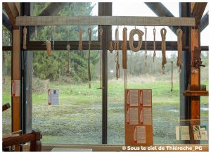 Ensemble de cordes et de nœuds suspendus à une large poutre au musée de la vie rurale et forestière de Saint-Michel. Les ficelles, cordages, échantillons torsadés et boucles servent à illustrer les techniques traditionnelles de la corderie. À l’arrière-plan, une grande baie vitrée ouvre sur un paysage boisé de la Thiérache. En bas, des panneaux explicatifs présentent l’histoire et les usages des différents nœuds.
