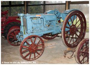 Tracteur agricole ancien exposé au musée de la vie rurale et forestière de Saint-Michel. La machine, peinte en bleu, possède de grandes roues métalliques rouges cerclées de fer, typiques des premiers modèles du XXᵉ siècle. Le moteur et la mécanique sont visibles en façade, témoignant des débuts de la motorisation agricole en Thiérache. En arrière-plan, on distingue un second tracteur rouge.