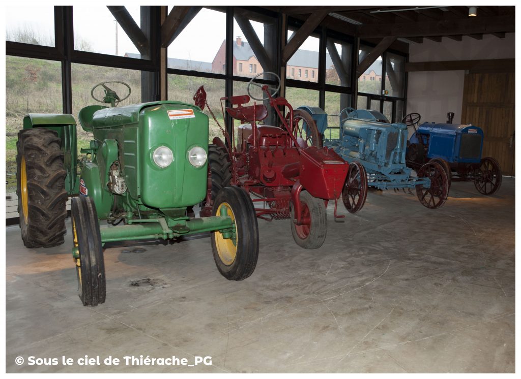 Alignement de tracteurs anciens verts, rouges et bleus exposés au musée de la vie rurale et forestière de Saint-Michel, devant une grande baie vitrée donnant sur la campagne thiérachienne.