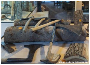 Haches et herminettes anciennes exposées au musée de la vie rurale et forestière de Saint-Michel. Les outils, posés sur des bûches de bois, montrent les formes variées des lames utilisées autrefois par les bûcherons et les charpentiers. La scène met en valeur le travail traditionnel du bois en Thiérache.