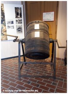 Grande baratte à beurre en bois montée sur un support métallique au musée de la vie rurale et forestière de Saint-Michel. Le tonneau cylindrique, équipé de poignées latérales permettant de le faire pivoter manuellement, évoque les anciennes techniques de fabrication du beurre en Thiérache. En arrière-plan, panneaux explicatifs et photographies d’époque.