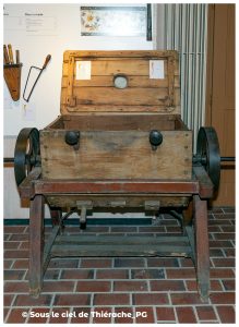 Grande baratte à beurre en bois exposée au musée de la vie rurale et forestière de Saint-Michel. L’appareil, monté sur un châssis en bois et métal, possède un large coffre ouvert et deux volants latéraux permettant d’actionner le mécanisme. À l’arrière-plan, un panneau présente des outils et informations sur les races bovines. L’ensemble illustre les anciennes techniques de fabrication du beurre en Thiérache.
