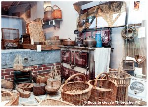 Reconstitution d’une cuisine et d’un atelier de vannerie au musée de la vie rurale et forestière de Saint-Michel. Au centre, une cuisinière ancienne ornée de motifs rouges est entourée d’ustensiles en métal, de pots et d’une cafetière bleue. Tout autour, de nombreux paniers, moules, outils et objets en osier témoignent du savoir-faire des vanniers. Des étagères présentent d’autres paniers et accessoires domestiques, composant une scène chaleureuse du quotidien rural en Thiérache.
