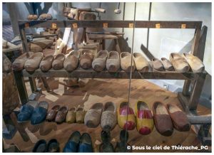 Présentoir de sabots en bois au musée de la vie rurale et forestière de Saint-Michel. Les étagères en bois accueillent des dizaines de sabots à différents stades de fabrication : bruts, semi-taillés, sculptés ou décorés. Certains modèles sont peints en couleurs vives, d’autres conservent l’aspect naturel du bois. En arrière-plan, des outils de sabotier complètent la reconstitution de cet artisanat traditionnel de Thiérache.