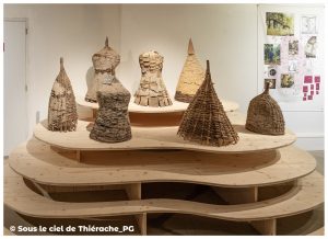 Ensemble de ruches traditionnelles exposées au musée de la vie rurale et forestière de Saint-Michel. Les ruches, de formes coniques ou arrondies, sont fabriquées en écorce, en torchis ou en osier tressé. Elles sont disposées sur un présentoir en bois aux plateaux ondulés. À l’arrière-plan, un panneau explicatif présente des photographies et schémas sur les techniques anciennes d’apiculture.