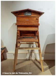 Ruche en bois de montagne exposée au musée de la vie rurale et forestière de Saint-Michel. La ruche, au toit en pente et à la façade ouvrante, repose sur un support en bois clair. Une petite ardoise indique son nom : « ruche ardente de montagne ». L’ensemble illustre les techniques d’apiculture traditionnelle.