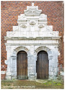 ue rapprochée du portail Renaissance de l’église fortifiée Saint-Médard de Parfondeval. L’entrée est composée de deux portes en bois massif, chacune encadrée par des arcs en plein cintre sculptés dans la pierre blanche. Le décor présente des pilastres, des chapiteaux ornés et des motifs Renaissance finement travaillés. Au-dessus, un fronton à plusieurs niveaux, richement sculpté de volutes et de niches, se détache sur le mur de brique rouge typique de la Thiérache, illustrant le contraste entre la robustesse défensive de l’édifice et l’élégance décorative du portail.