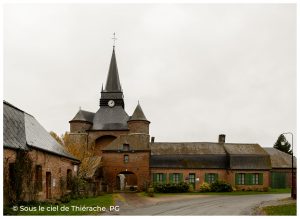 Vue d’ensemble de l’église fortifiée Saint-Médard de Parfondeval, construite en brique rouge, avec son imposant donjon trapu et de plan carré, encadré de deux tours circulaires, surmonté d’un clocher central coiffé d’une flèche et d’une horloge. L’édifice, typique de l’architecture fortifiée thiérachienne, domine la place du village, entouré de bâtiments communaux en brique et de verdure, sous un ciel couvert.