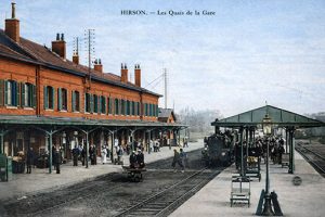 Carte postale ancienne colorisée montrant les quais de la gare d’Hirson au début du XXᵉ siècle : bâtiment voyageurs en briques avec marquise, locomotive à vapeur à l’arrêt, voyageurs et bagages sur les quais, lampadaires et mobilier ferroviaire d’époque.