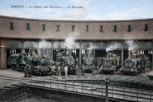 Carte postale ancienne colorisée montrant la rotonde du dépôt des machines de la gare d’Hirson : plusieurs locomotives à vapeur alignées dans le demi-cercle, cheminots rassemblés sur le pont tournant, fumées s’échappant des machines, scène emblématique de l’activité ferroviaire au début du XXᵉ siècle.