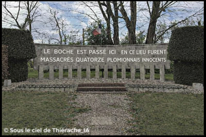 Monument commémoratif à Plomion : un mur de pierre portant l’inscription « Le Bosche est passé ici – en ce lieu furent massacrés quatorze Plomionnais innocents », devant une rangée de stèles, avec un drapeau français en arrière-plan et un chemin de gravier menant au mémorial.