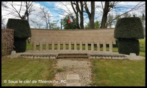 Monument des Martyrs à Plomion, mémorial en pierre dressé dans un espace paysager, composé d’un mur commémoratif gravé, de piliers alignés et encadré par des haies taillées, avec un drapeau français visible à l’arrière-plan.