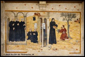 Peinture murale religieuse restaurée représentant deux scènes médiévales : à gauche, des moines vêtus de robes noires se tiennent et prient sous des arcades, tandis que deux autres sont agenouillés au premier plan. Au centre, un élément architectural avec porte et fenêtres sépare la composition. À droite, une religieuse en habit noir remet du pain à un homme agenouillé près d’un arbre, dans un paysage vallonné aux tons ocres, avec des bâtiments au loin. L’ensemble présente des couleurs ravivées tout en conservant l’aspect ancien de la fresque.