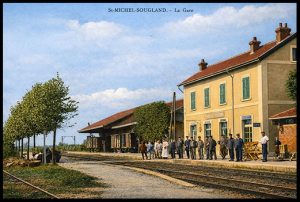 Carte postale ancienne colorisée de la gare de Saint-Michel-Sougland : bâtiment voyageurs aux façades claires et volets verts bordant les voies ferrées, quais arborés et groupe de voyageurs et de cheminots rassemblés devant la gare sous un ciel bleu