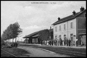 Carte postale ancienne en noir et blanc de la gare de Saint-Michel-Sougland : bâtiment voyageurs à deux niveaux le long des voies ferrées, quais bordés d’arbres, avec un groupe de voyageurs et de cheminots rassemblés devant la gare.