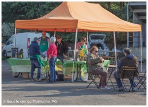Stand de marché local sous un barnum orange, où plusieurs producteurs accueillent des visiteurs autour de tables couvertes de nappes vertes. Des clients échangent avec les vendeurs tandis que d’autres sont assis à une petite table en train de consommer, dans une ambiance conviviale de marché de plein air. En arrière-plan, des véhicules utilitaires et des arbres encadrent la scène.