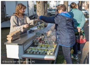 Vente sur un marché de plein air : une productrice tend un produit à une cliente au-dessus d’une vitrine réfrigérée remplie de yaourts et desserts. D’autres visiteurs attendent à proximité, sac à la main, dans une ambiance simple et conviviale typique d’un marché local en Thiérache.