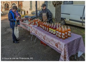 Stand de marché proposant des jus et produits du verger disposés en bouteilles alignées sur une table. Une cliente consulte son porte-monnaie avant un achat, tandis que le producteur se tient derrière l’étal. À côté, des œufs sont présentés en plateaux, dans une ambiance simple et authentique de marché de village en Thiérache.