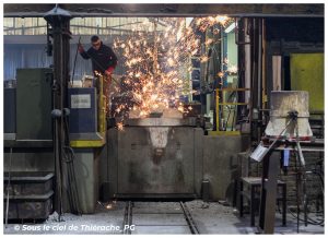 Un ouvrier de fonderie travaille au-dessus d’un moule de métal dans un atelier industriel, tandis qu’une pluie d’étincelles jaillit lors de l’opération de coulée ou de nettoyage de la pièce, au cœur des Fonderies de Sougland à Saint-Michel en Thiérache. L’atelier est entouré de machines, d’outils et de structures métalliques typiques d’un site de production de fonte