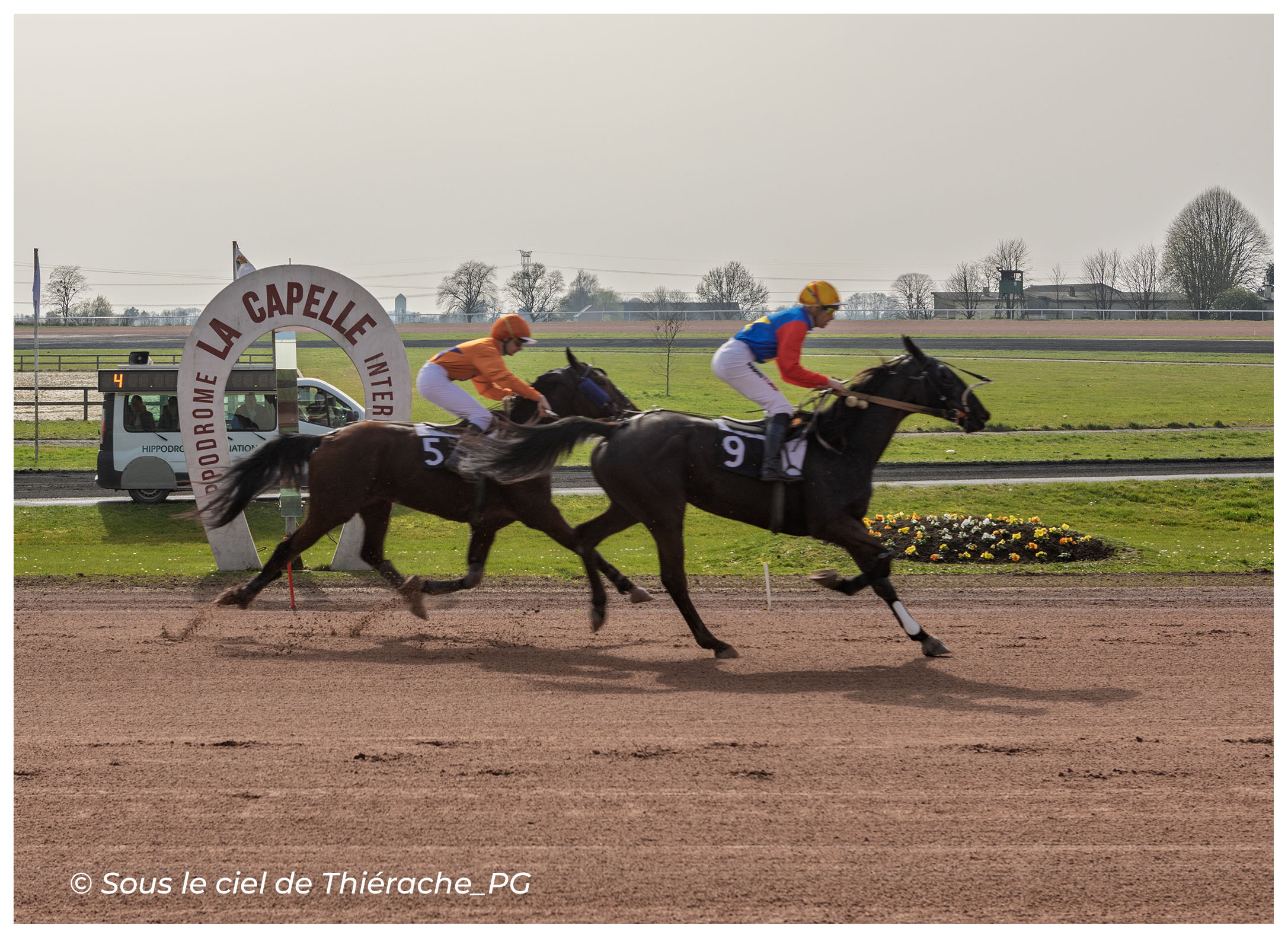 Course hippique à l’hippodrome international de La Capelle en Thiérache. Deux chevaux numérotés 5 et 9 galopent à vive allure sur la piste de sable tandis que leurs jockeys, vêtus de casaques colorées, se disputent la position devant l’arche marquée “Hippodrome La Capelle”.