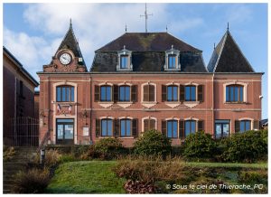 Façade de la mairie de Saint-Michel-en-Thiérache, bâtiment communal symétrique en briques rosées avec volets en bois, horloge et inscription « Ville de Saint-Michel », surmonté d’un toit d’ardoise à lucarnes et encadré de deux pavillons pointus, devant un petit jardin sous un ciel nuageux.