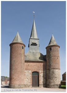 Église fortifiée de La Bouteille en Thiérache, façade en briques rouges avec deux tours cylindriques défensives et clocher central en ardoise, sous ciel bleu.