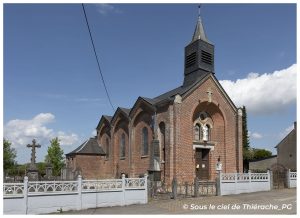 Vue de l’église de Sommeron en Thiérache et de son monument aux morts, au cœur du village, avec architecture en briques rouges typique de la Thiérache et cimetière communal sous un ciel d’été.