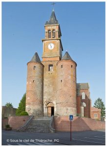Façade imposante d’une église fortifiée en brique et pierre typique de la Thiérache, photographiée sous un ciel bleu. L’entrée principale est encadrée par deux grandes tours cylindriques défensives coiffées de toitures coniques en ardoise. Au-dessus s’élève un clocher-porche surmonté d’une flèche élancée et d’une horloge visible. Un large escalier mène au portail roman, tandis que les volumes massifs de l’édifice témoignent de son rôle à la fois religieux et défensif. Photo prise en lumière douce mettant en valeur les détails architecturaux et les traces du temps sur les murs.