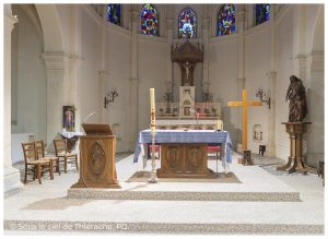 Vue du maître-autel d’une église fortifiée de Thiérache photographié depuis le centre du chœur. Au premier plan, un autel en bois sculpté recouvert d’une nappe bleue et blanche est entouré d’un pupitre de lecture, d’un grand cierge pascal et d’une grande croix en bois. À l’arrière-plan, le retable et le tabernacle sont surmontés d’un crucifix placé sous un élégant baldaquin en bois sculpté. De hauts vitraux colorés illuminent l’espace et mettent en valeur les colonnes élancées ainsi que l’architecture gothique sobre et harmonieuse