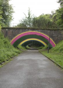 Vue d’ensemble du pont de la Ruelle Thiébault à Vervins après sa métamorphose artistique. Les arches aux couleurs vives – vert, rose, jaune et violet – se détachent dans un écrin de verdure et annoncent l’univers poétique qui se dévoile sous l’ouvrage. Cette fresque monumentale réalisée par Dorothée Ossart transforme un simple passage en une véritable porte d’entrée vers un jardin imaginaire inspiré des souvenirs d’enfance de l’artiste.