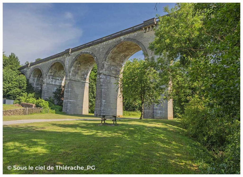 Vue en contre-plongée du viaduc d'Ohis, imposant ouvrage d'art ferroviaire en pierre composé de grandes arches. Entouré d'arbres et d'une prairie verdoyante, il est aujourd'hui un site emblématique de l'Axe Vert de la Thiérache.