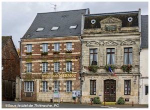Façade du musée Jean Mermoz et de la mairie d'Aubenton, sur la place centrale du village de Thiérache. L'édifice en briques et pierres, typique de l'architecture locale, côtoie un élégant bâtiment en pierre de taille orné de balcons fleuris et du drapeau français.